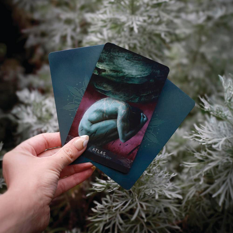 Hand holding mystical tarot card 'Atlas' with ethereal artwork, surrounded by soft foliage for spiritual reading.