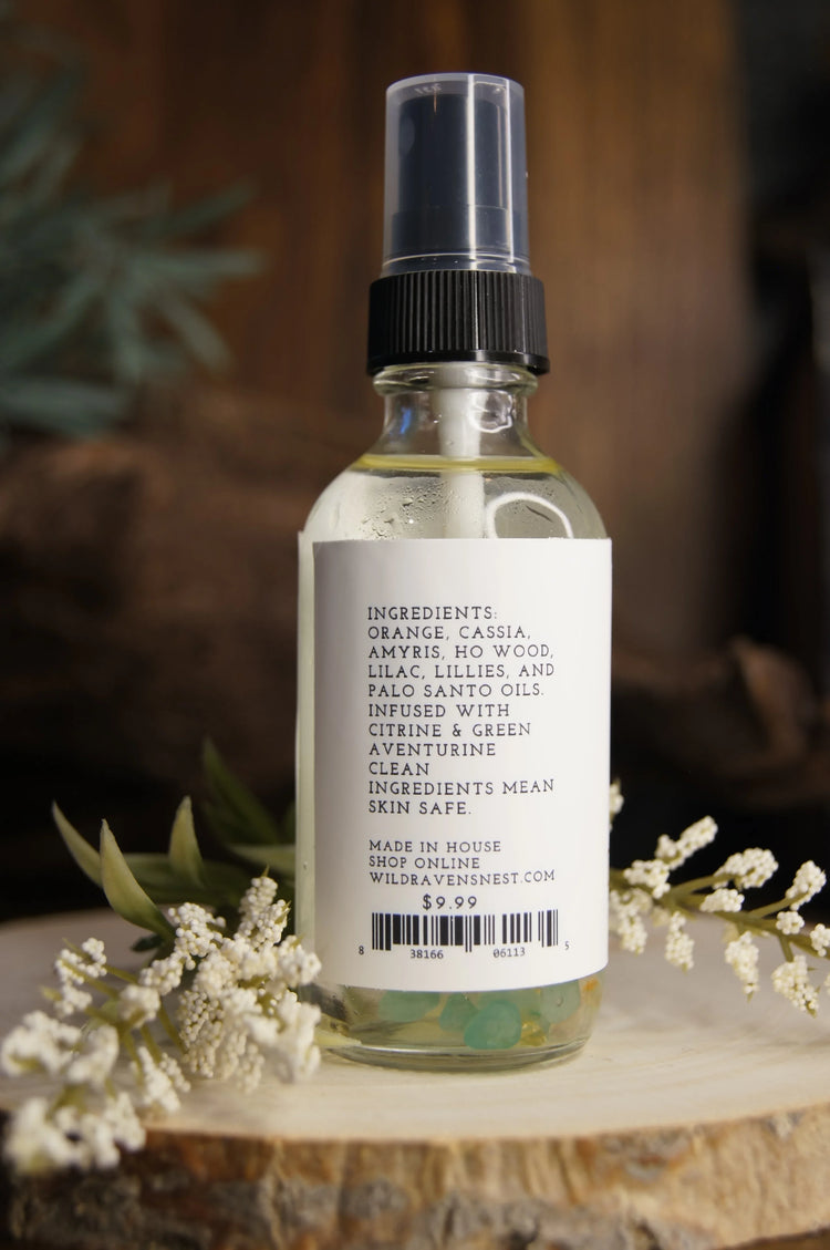 Glass spray bottle with botanical oil ingredients and crystals, placed on wood with flowers.