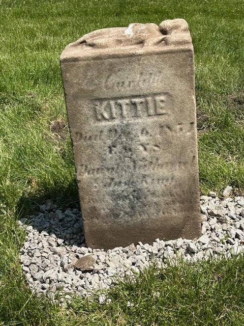 Old weathered gravestone labeled KITTIE on grass with stones at the base, in sunlight.
