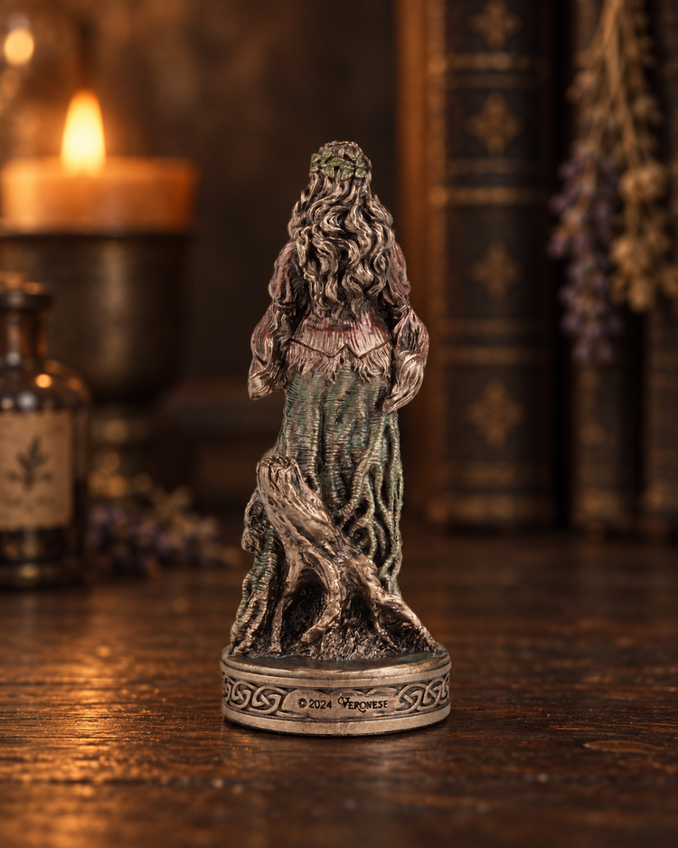 A fantasy figurine of Danu with long wavy hair and a tree-like body, standing on a circular base, photographed on a wooden surface with a blurred candle and books in the background.