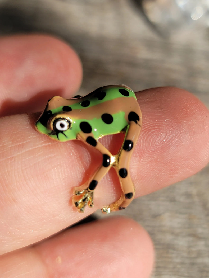 Close-up of a tiny green and tan spotted frog figurine with gold accents on a fingertip