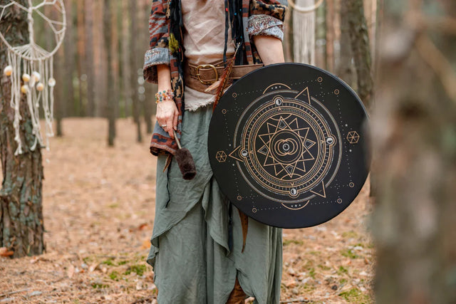 Person in boho clothing holding a large shamanic drum with geometric design in a forest setting.