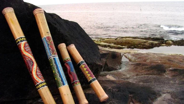 Four decorated bamboo rainsticks with colorful patterns rest on rocks by the ocean shore.