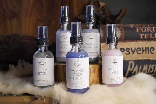 Five spiritual aura and room sprays with crystal accents, displayed on fur and wood by metaphysical books.