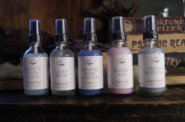 Five aura and room sprays with spiritual themes, arranged on a wooden shelf with metaphysical decor.