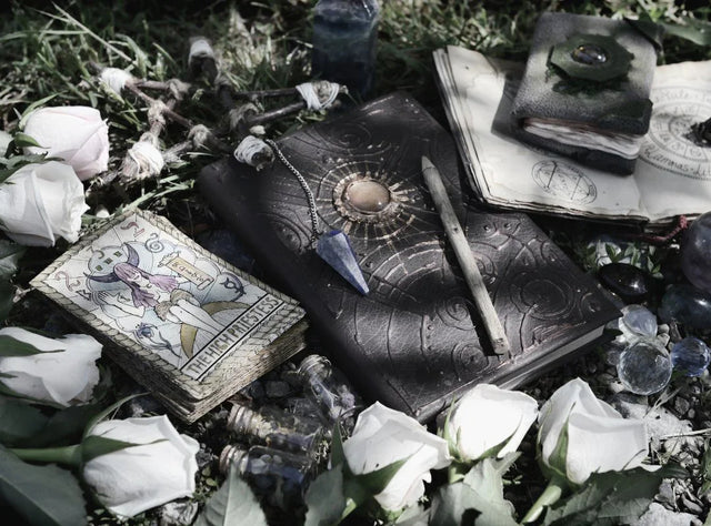 Tarot cards, pendulum, crystals, spell book, and white roses on grass outdoors