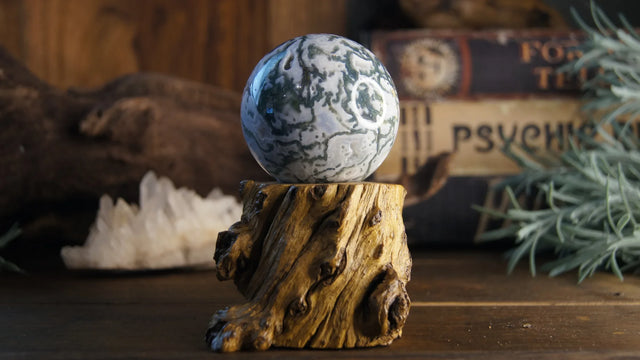 Moss agate crystal sphere on wooden stand with books, greenery, and quartz in background