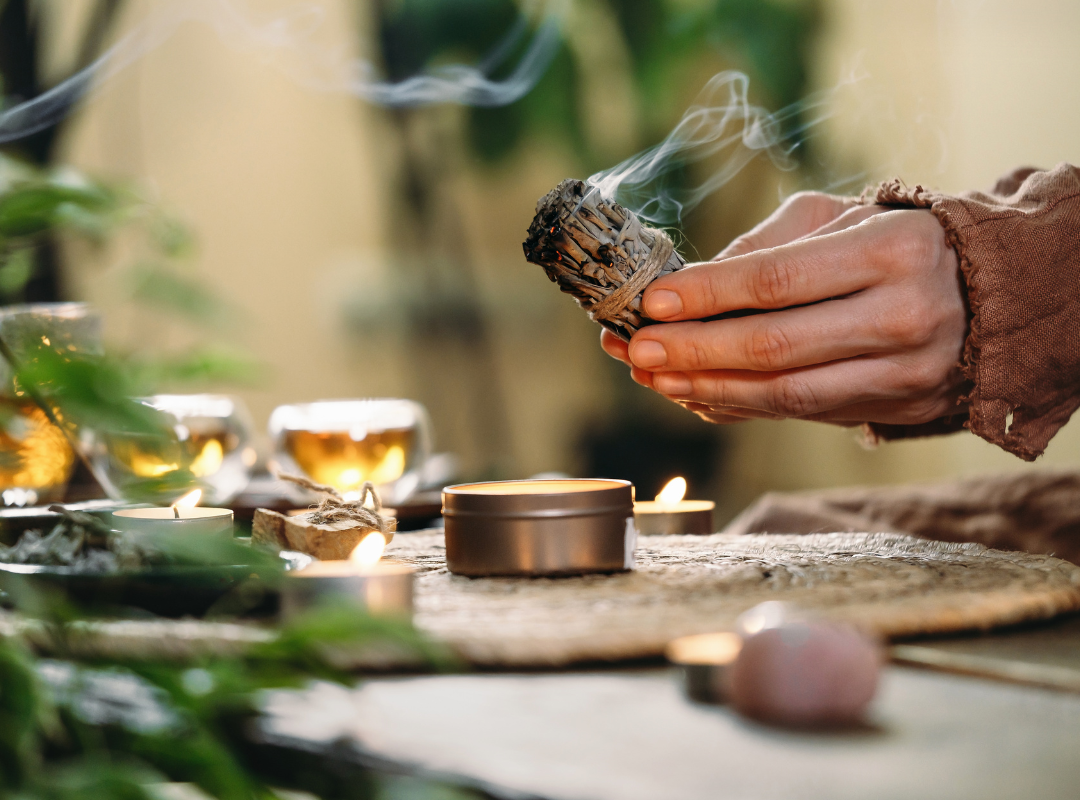 Hand holding smudge stick near candles, crystals, and tea in a calming spiritual setting
