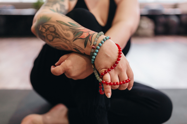 Tattooed arm with gemstone beaded bracelets, seated in a yoga pose on a mat