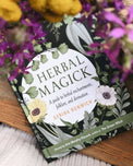 Herbal Magick book with botanical cover on table surrounded by purple flowers, metaphysical decor