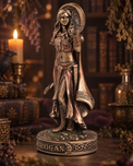 A detailed figurine of the goddess Morrigan standing on a circular base inscribed with 'MORRIGAN'. The statue features a weathered metallic finish with bronze, copper, and dark brown tones, set against a dark, atmospheric background of old books, a lit candle, and dried botanicals.