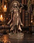 A bronze-finished figurine of the Celtic goddess Morrigan standing on a decorative base labeled 'MORRIGAN', with a raven perched on her raised hand, photographed in a dark, atmospheric setting with candles, books, and dried herbs.