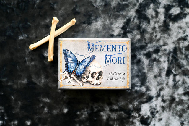 Memento Mori oracle deck box with blue butterfly, skull, and crossed bones on marbled surface