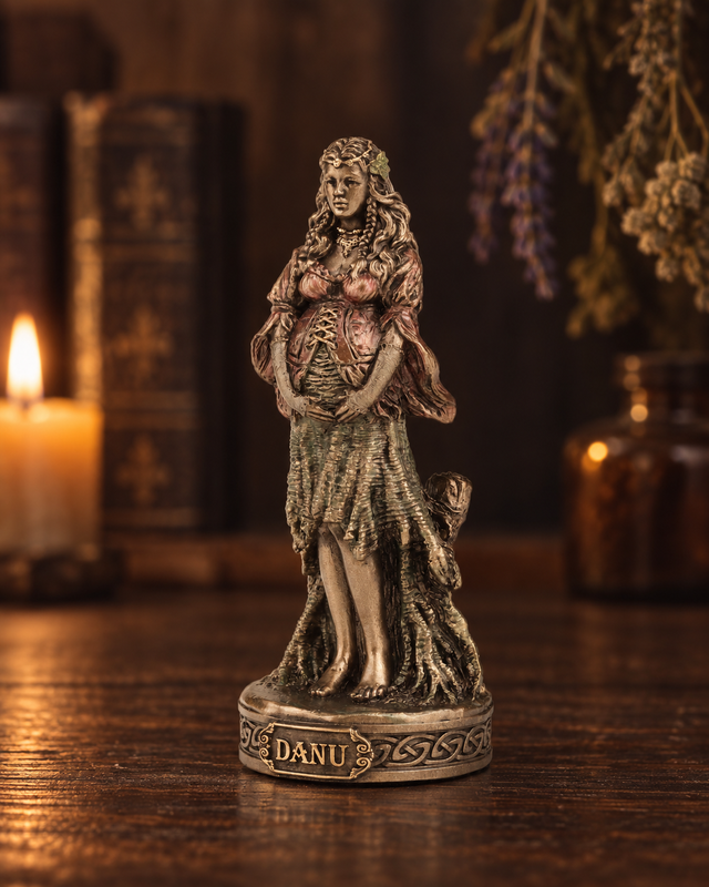 A small bronze-finished figurine of a pregnant goddess standing on a base labeled 'DANU', displayed on a wooden table with a blurred background containing a lit candle, books, and dried lavender.