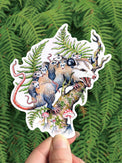 Opossum Queen vinyl sticker, an opossum among mushrooms and forest greens, held against ferns